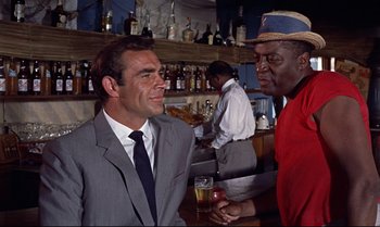 Movie still from “Dr. No” (1962), directed by Terence Young – A man in a gray suit is talking to another man in a red shirt; Medium shot, Over the shoulder angle