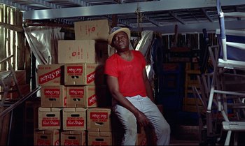 Movie still from “Dr. No” (1962), directed by Terence Young – A man sitting on a pile of boxes in a room; Medium shot, High angle
