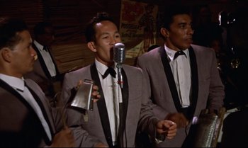 Movie still from “Dr. No” (1962), directed by Terence Young – A man in a gray suit is singing into a microphone with another man in a gray suit; Medium shot, High angle
