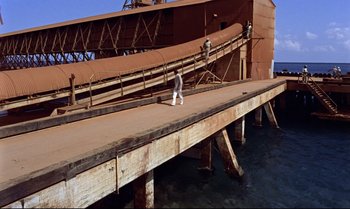 Movie still from “Dr. No” (1962), directed by Terence Young – A man walking across a wooden bridge near water; Extreme Wide shot, High angle