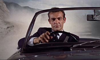 Movie still from “Dr. No” (1962), directed by Terence Young – A man sitting in the driver's seat of a car while holding a steering wheel; Close Up shot, Low angle
