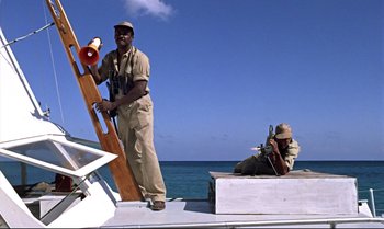 Movie still from “Dr. No” (1962), directed by Terence Young – A man standing on top of a boat next to another man sitting on a boat; Wide shot, Low angle
