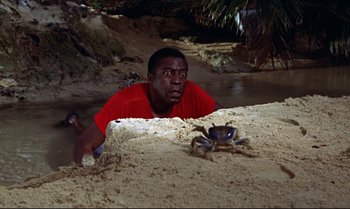 Movie still from “Dr. No” (1962), directed by Terence Young – A man in a red t - shirt is looking at a crab on the beach; Medium shot, High angle