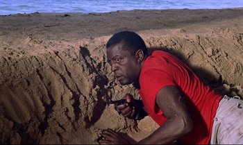Movie still from “Dr. No” (1962), directed by Terence Young – A man in a red t - shirt leaning over a sandy beach; Medium shot, High angle