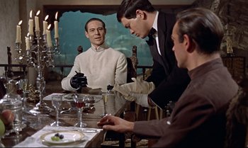 Movie still from “Dr. No” (1962), directed by Terence Young – A group of people sitting at a table with a plate; Medium shot, Over the shoulder angle