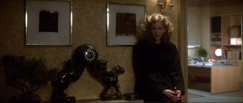 Movie still from “Dressed to Kill” (1980), directed by Brian De Palma – A woman sitting in front of a wall with a clock and picture on the wall; Medium shot, Low angle