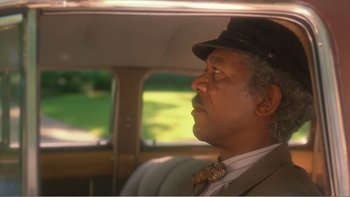 Movie still from “Driving Miss Daisy” (1989), directed by Bruce Beresford – A man sitting in the back seat of a car wearing a hat; Close Up shot, Over the shoulder angle
