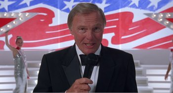 Movie still from “Drop Dead Gorgeous” (1999), directed by Michael Patrick Jann – An older man holding a microphone in front of an american flag; Close Up shot, High angle