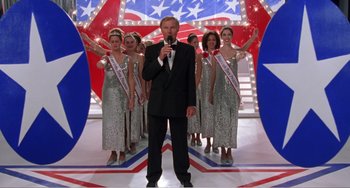 Movie still from “Drop Dead Gorgeous” (1999), directed by Michael Patrick Jann – A man in a suit is standing in front of a group of women in silver sequined dresses; Wide shot, Low angle