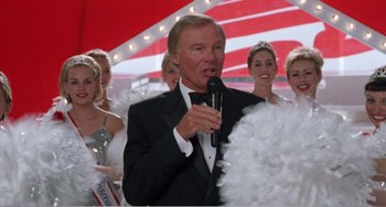 Movie still from “Drop Dead Gorgeous” (1999), directed by Michael Patrick Jann – A man in a tuxedo is holding a microphone; Medium shot, Low angle