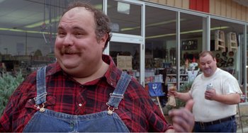 Movie still from “Drop Dead Gorgeous” (1999), directed by Michael Patrick Jann – A man in a plaid shirt and blue overalls standing in front of a store; Medium shot, Low angle