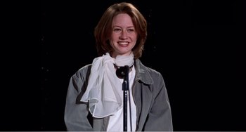 Movie still from “Drop Dead Gorgeous” (1999), directed by Michael Patrick Jann – A woman standing in front of a microphone wearing a white scarf; Medium shot, Low angle