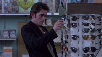 Movie still from “Drugstore Cowboy” (1989), directed by Gus Van Sant – A man in black jacket looking at sunglasses in a store; Medium shot, Over the shoulder angle