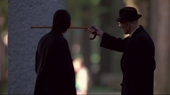 Movie still from “Drugstore Cowboy” (1989), directed by Gus Van Sant – A man in a hat is holding a cane over another man's head; Medium shot, Over the shoulder angle