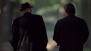 Movie still from “Drugstore Cowboy” (1989), directed by Gus Van Sant – Two men in black suits and a hat are walking; Medium shot, Over the shoulder angle