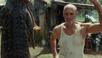 Movie still from “The Paperboy” (2012), directed by Lee Daniels – A bald man in a white tank top is standing next to an alligator; Medium shot, Low angle