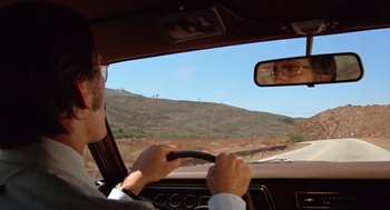 Movie still from “Duel” (1971), directed by Steven Spielberg – A man driving a car on a road with hills in the background; Wide shot, Over the shoulder angle