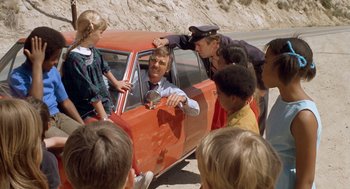 Movie still from “Duel” (1971), directed by Steven Spielberg – A group of people standing next to an orange car on the side of the road; Wide shot, High angle