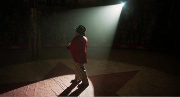 Movie still from “Dumbo” (2019), directed by Tim Burton – A man in a red coat and a black hat is standing in the spotlight; Wide shot, Low angle