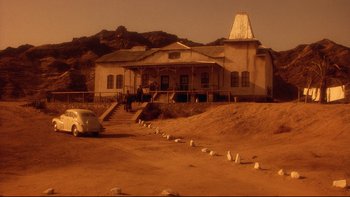 Movie still from “Dust Devil” (1992), directed by Richard Stanley – An old car is parked in front of an abandoned building; Extreme Wide shot, Low angle