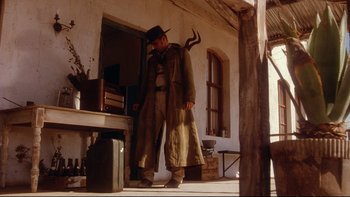 Movie still from “Dust Devil” (1992), directed by Richard Stanley – A man with a horned hat standing in front of a building; Wide shot, Low angle