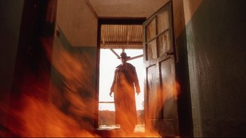 Movie still from “Dust Devil” (1992), directed by Richard Stanley – A man standing in front of a door with fire coming out of it; Wide shot, Low angle