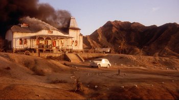 Movie still from “Dust Devil” (1992), directed by Richard Stanley – A white van parked in front of a house on fire; Extreme Wide shot, High angle