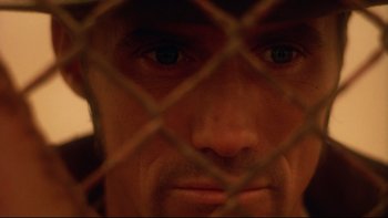 Movie still from “Dust Devil” (1992), directed by Richard Stanley – A man's face behind a chain link fence; Extreme Close Up shot, Overhead angle