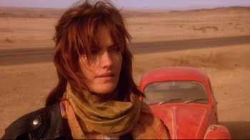 Movie still from “Dust Devil” (1992), directed by Richard Stanley – A woman in a scarf standing in front of a red car; Close Up shot, Low angle