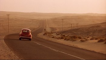 Movie still from “Dust Devil” (1992), directed by Richard Stanley – An old red car driving down the middle of a desert road; Extreme Wide shot, High angle