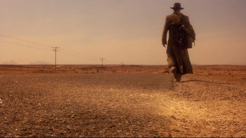 Movie still from “Dust Devil” (1992), directed by Richard Stanley – A man walking across a dirt road in the middle of the desert; Wide shot, Low angle