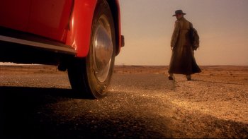 Movie still from “Dust Devil” (1992), directed by Richard Stanley – A man walking down the road next to a red car; Wide shot, Low angle