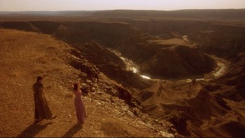 Movie still from “Dust Devil” (1992), directed by Richard Stanley – A woman standing on top of a hill looking at a valley; Extreme Wide shot, High angle