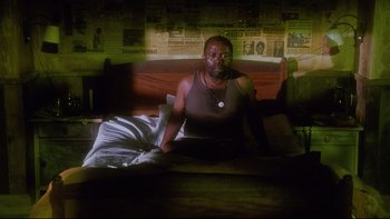 Movie still from “Dust Devil” (1992), directed by Richard Stanley – A man sitting on top of a bed in a room; Medium shot, High angle