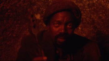 Movie still from “Dust Devil” (1992), directed by Richard Stanley – A man holding a guitar in front of a wall; Close Up shot, High angle