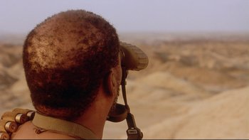 Movie still from “Dust Devil” (1992), directed by Richard Stanley – A man looking through binoculars in the desert; Close Up shot, Over the shoulder angle