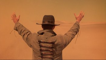 Movie still from “Dust Devil” (1992), directed by Richard Stanley – A man in a cowboy hat and jacket with his arms outstreched; Medium shot, Low angle