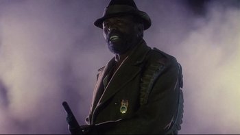 Movie still from “Dust Devil” (1992), directed by Richard Stanley – A man in a brown jacket holding a gun and wearing a hat; Close Up shot, Low angle