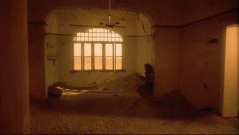 Movie still from “Dust Devil” (1992), directed by Richard Stanley – A person sitting in a room with a window in the middle of the room; Extreme Wide shot, Low angle