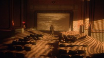 Movie still from “Dust Devil” (1992), directed by Richard Stanley – A man standing in front of a projection screen in an auditorium; Extreme Wide shot, High angle