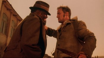 Movie still from “Dust Devil” (1992), directed by Richard Stanley – Two men in suits and a hat one of which has a knife in his other's hand; Medium shot, Over the shoulder angle