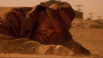 Movie still from “Dust Devil” (1992), directed by Richard Stanley – A person laying in the sand wearing a hat; Close Up shot, Low angle