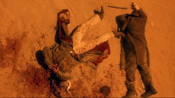 Movie still from “Dust Devil” (1992), directed by Richard Stanley – A man is lying on the ground with a knife in his hand; Wide shot, Overhead angle
