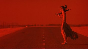 Movie still from “Dust Devil” (1992), directed by Richard Stanley – A woman standing on the side of a road holding a cell phone; Wide shot, Low angle