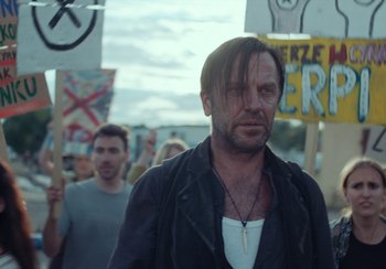 Movie still from “EO” (2022), directed by Jerzy Skolimowski – A man in a black jacket is standing in front of a group of people; Close Up shot, Low angle