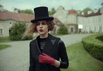 Movie still from “EO” (2022), directed by Jerzy Skolimowski – A woman wearing a top hat and gloves in front of a house; Medium shot, Low angle
