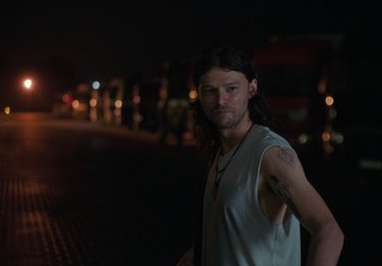 Movie still from “EO” (2022), directed by Jerzy Skolimowski – A man with long black hair standing on the side of a road; Medium shot, Low angle