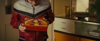 Movie still from “Eddie the Eagle” (2015), directed by Dexter Fletcher – A person holding a box in a room; Medium shot, High angle