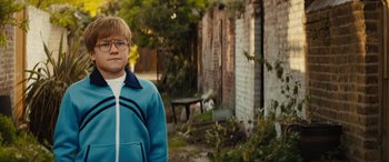 Movie still from “Eddie the Eagle” (2015), directed by Dexter Fletcher – A young man standing in front of a brick building; Close Up shot, Over the shoulder angle