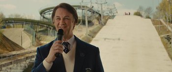 Movie still from “Eddie the Eagle” (2015), directed by Dexter Fletcher – A man holding a microphone in front of a ski lift; Medium shot, Low angle